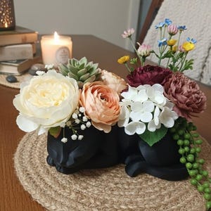 May include: A black cat-shaped planter filled with an assortment of colourful faux flowers. The arrangement includes cream, peach, burgundy, and white blooms, with green accents. The planter sits on a round, woven jute placemat.