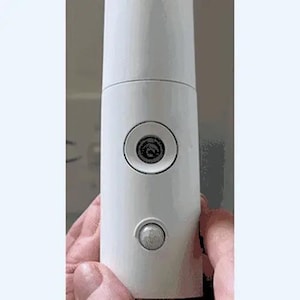 May include: A white cylindrical electronic device featuring a circular power button with a central icon and a smaller, translucent circular motion sensor positioned below it on the smooth, matte-finished exterior surface of the main housing.