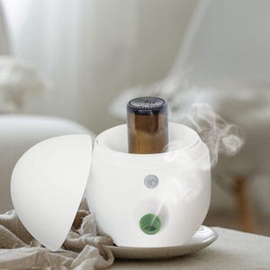 May include: White diffuser with a round lid and a small green vent. A brown glass bottle is placed inside the diffuser. Steam is coming out of the diffuser.