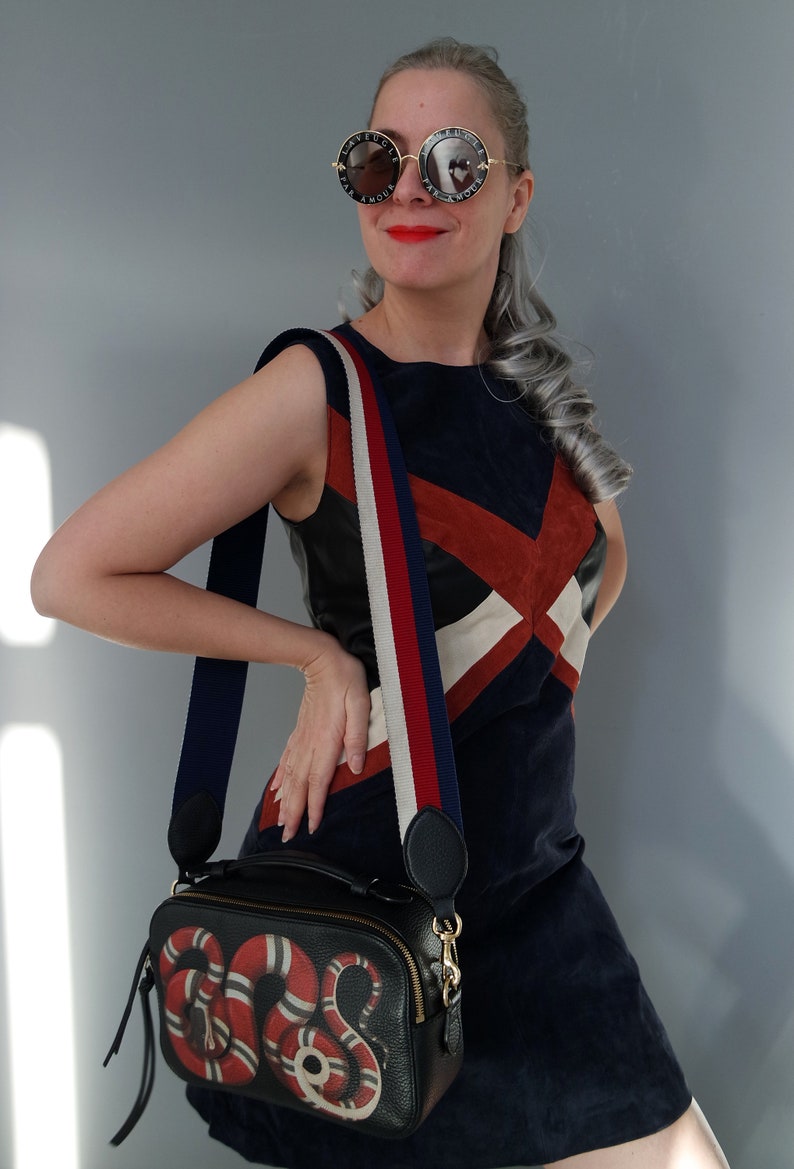 K&ouml;nnte beinhalten: Schwarze Leder-Umh&auml;ngetasche mit einem rot-wei&szlig;en Schlangenmuster. Die Tasche hat einen gestreiften Schulterriemen in Marineblau, Rot und Wei&szlig;. Die Frau tr&auml;gt ein marineblaues Kleid mit geometrischem Design und runde Sonnenbrille.