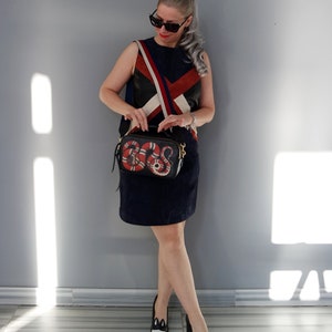 K&ouml;nnte beinhalten: Eine Frau tr&auml;gt ein marineblaues Kleid, eine Sonnenbrille und schwarz-wei&szlig;e Schuhe. Sie h&auml;lt eine schwarze Umh&auml;ngetasche mit einem rot-wei&szlig;en Schlangenmuster. Das Outfit umfasst ein Oberteil mit einem rot-wei&szlig;-blauen Design.
