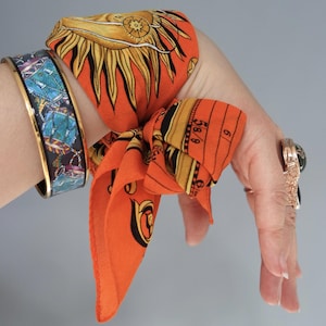 May include: An orange silk scarf featuring a sun and measurement design is tied around a wrist. A blue and gold cuff bracelet and a rose gold ring with a black stone are also visible. The jewellery is set against a grey background.