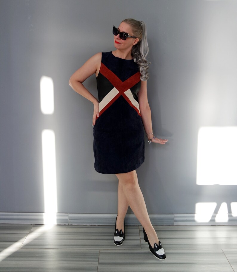 K&ouml;nnte beinhalten: Ein &auml;rmelloses marineblaues Kleid mit einem geometrischen Design in Rot, Schwarz und Creme. Das Outfit wird durch schwarz-wei&szlig;e Schuhe und Sonnenbrille erg&auml;nzt. Die Frau lehnt an einer grauen Wand.