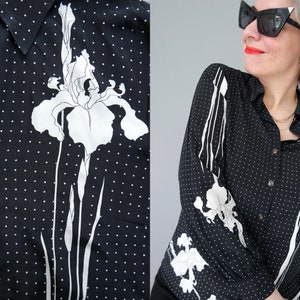 Op de afbeelding: Zwart overhemd met witte stippen en een witte irisbloemprint. Het shirt met lange mouwen heeft een kraag en een zak op de linkerborst. Het irisontwerp staat ook op de mouwen. Knoopsluiting.