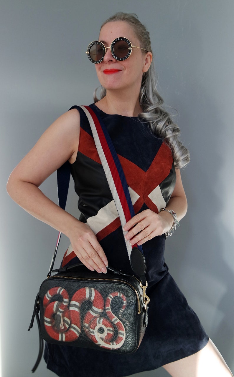 K&ouml;nnte beinhalten: Eine Frau tr&auml;gt eine runde Sonnenbrille mit Text, ein marineblaues und rotes Kleid und eine schwarze Umh&auml;ngetasche. Die Tasche hat ein rotes und wei&szlig;es Schlangenmuster. Der Riemen ist rot, wei&szlig; und blau gestreift.