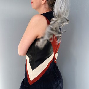 K&ouml;nnte beinhalten: Ein &auml;rmelloses, marineblaues Kleid mit einem Farbblock-Design mit schwarzen, wei&szlig;en und roten Akzenten. Das Kleid hat einen ausgestellten Rock und ist aus einem Wildleder-&auml;hnlichen Material gefertigt. Die Person tr&auml;gt eine Sonnenbrille.