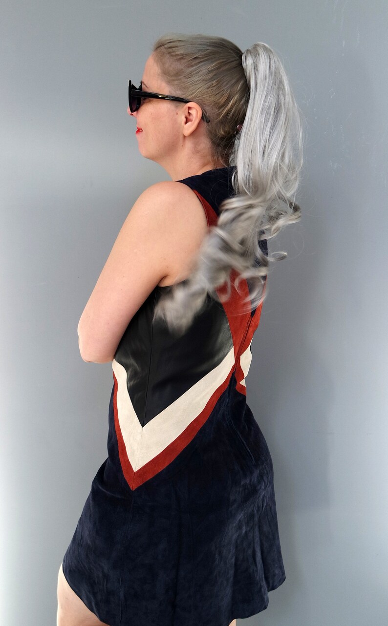 K&ouml;nnte beinhalten: Ein &auml;rmelloses, marineblaues Kleid mit einem Farbblock-Design mit schwarzen, wei&szlig;en und roten Akzenten. Das Kleid hat einen ausgestellten Rock und ist aus einem Wildleder-&auml;hnlichen Material gefertigt. Die Person tr&auml;gt eine Sonnenbrille.