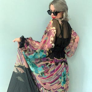 May include: A sheer black dress with a colourful patterned overlay. The overlay features a vibrant design with pink, purple, blue, and gold hues. The dress has long sleeves and a cinched waist, creating an elegant silhouette.