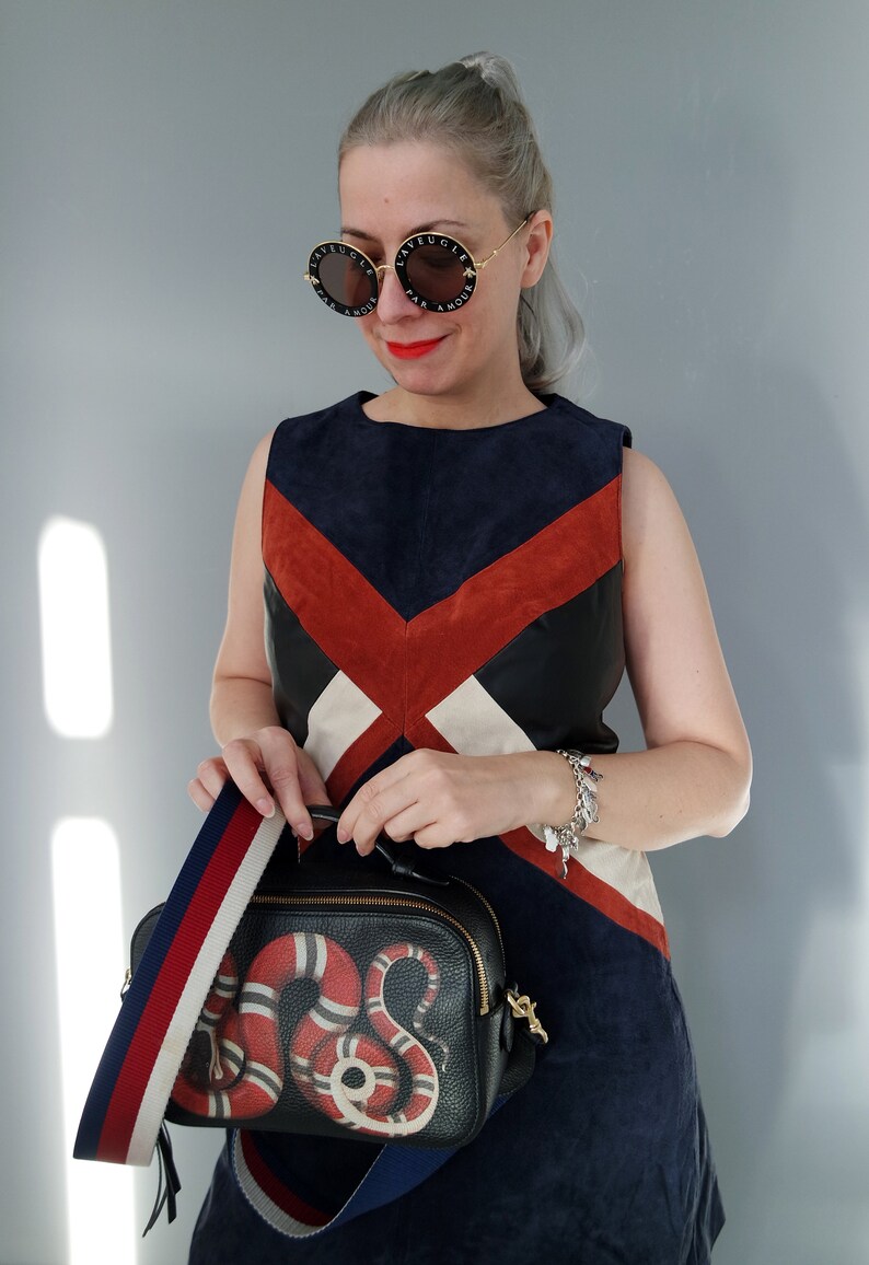 K&ouml;nnte beinhalten: Eine Frau tr&auml;gt ein farbblockiertes Kleid und runde Sonnenbrillen. Sie tr&auml;gt eine schwarze Lederhandtasche mit einem rot-wei&szlig;en Schlangenmuster und einem gestreiften Riemen. Die Sonnenbrille hat Text um die Gl&auml;ser.