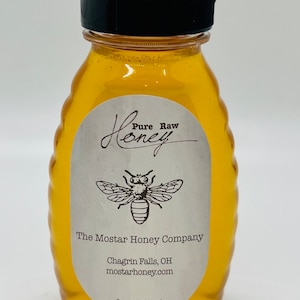 May include: A clear glass bottle with a black lid filled with honey. The label reads "Pure Raw Honey" with a bee illustration and "The Mostar Honey Company, Chagrin Falls, OH, mostarhoney.com, 8 oz. (227 g.)"