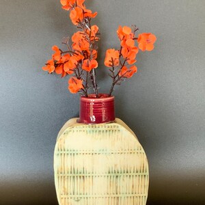 May include: A decorative ceramic vase with a textured, off-white body and a small, deep red top. The vase holds vibrant orange flowers with dark brown stems, creating a striking contrast. The vase has a unique, artistic design.