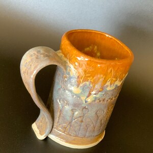 May include: Handmade ceramic mug with a unique, curved handle. The mug features a gradient of colors, including orange, blue, and brown. The exterior has a textured floral design. The interior is a warm orange color.