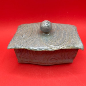 May include: A square ceramic trinket box with a lid. The box is a muted gray-green color with a textured, swirling pattern on the lid and sides. A small, round knob serves as a handle on the lid.