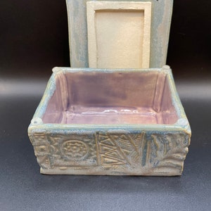 May include: A ceramic box with a light blue and green glaze. The box has a raised design on the outside and a purple interior. The lid is open and the box is empty.