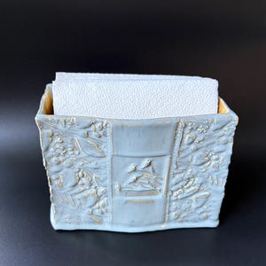 May include: A light blue ceramic napkin holder with a rectangular shape and a textured surface featuring embossed botanical patterns and a central panel depicting small birds. The holder contains a stack of white, textured paper napkins.