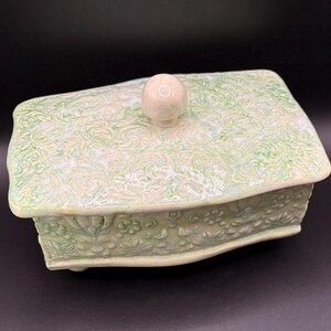May include: A rectangular ceramic box with a lid, featuring a light green and white floral pattern. The box has a rounded top and a small, round knob on the lid. The sides of the box are also decorated with floral designs.
