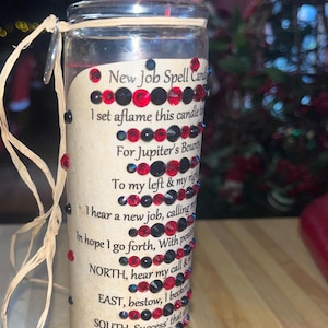May include: A glass candle jar with a beige label and red and black rhinestones. The label has the text "New Job Spell Candle" and a poem about finding a new job.