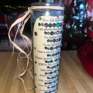 May include: A clear glass candle with a green and gold label that reads "New Job Spell Candle". The label is decorated with iridescent rhinestones. The candle is surrounded by a straw-like material.