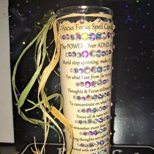 May include: A glass jar candle with a brown paper label that reads "Hocus Focus Spell Candle The POWER Over ADHD-ADD World stop spinning, make it slow For what I see from far to near Thoughts & Focus stillness To concentrate on what I see Focus all & nary not I remember more than I forget  Keeping thoughts are calm & relaxed with ease & no more Keep me paced from inside I'm in control without a doubt Thoughts & Focus stillness This Spell is Cast, so mote it be". The jar is decorated with iridescent rhinestones.