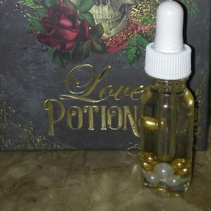 May include: A clear glass bottle with a white dropper top filled with a golden liquid and white pearls. The bottle is labeled "Love Potion" in gold lettering on a black background with a skull and roses.