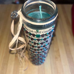 May include: A teal blue candle with a white wick in a clear glass jar. The jar is decorated with a brown and green ribbon and rhinestones. The label on the jar reads "New Job Spell Candle" and includes a short poem about finding a new job.
