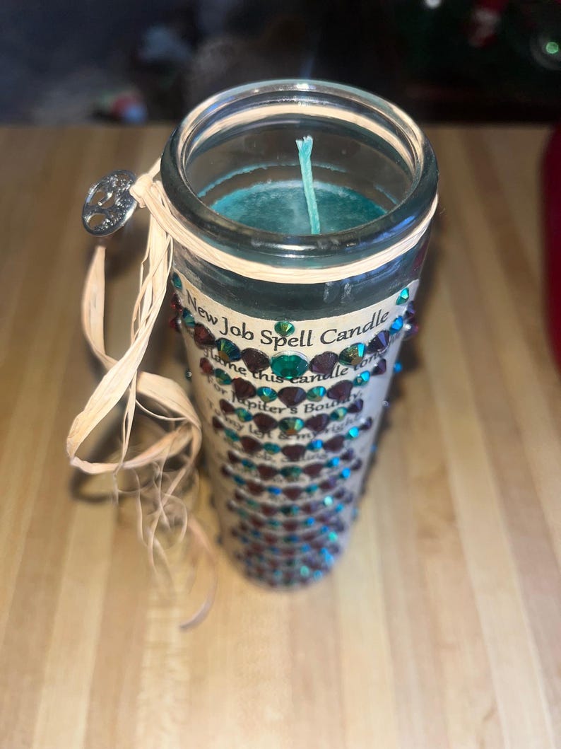 May include: A teal blue candle with a white wick in a clear glass jar. The jar is decorated with a brown and green ribbon and rhinestones. The label on the jar reads "New Job Spell Candle" and includes a short poem about finding a new job.