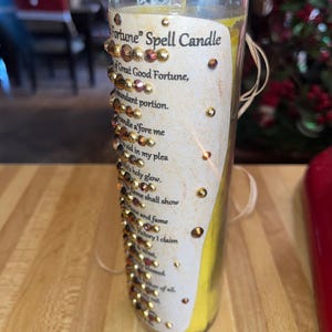 K&ouml;nnte beinhalten: Eine gelbe Zauberkerze in einem durchsichtigen Glasgef&auml;&szlig; mit einem wei&szlig;en Etikett, auf dem "Fortune" Spell Candle steht. Das Etikett ist mit goldenem Glitzer und Strasssteinen verziert und lautet "Great Good Fortune, abundant portion. Candle a'fore me to aid in my plea. Holy glow. Flame shall show and fume. Victory I claim. Blessed of all."
