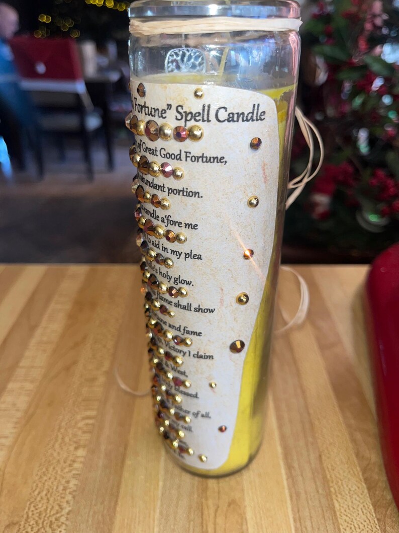 K&ouml;nnte beinhalten: Eine gelbe Zauberkerze in einem durchsichtigen Glasgef&auml;&szlig; mit einem wei&szlig;en Etikett, auf dem "Fortune" Spell Candle steht. Das Etikett ist mit goldenem Glitzer und Strasssteinen verziert und lautet "Great Good Fortune, abundant portion. Candle a'fore me to aid in my plea. Holy glow. Flame shall show and fume. Victory I claim. Blessed of all."