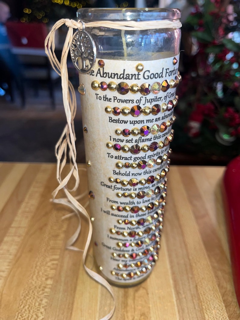 K&ouml;nnte beinhalten: Ein Glas-Kerzengef&auml;&szlig; mit einem beigefarbenen Etikett und goldenen Glitzerakzenten. Das Etikett tr&auml;gt den Text "The Abundant Good Fortune" und ein Gedicht &uuml;ber das Anziehen von Gl&uuml;ck.