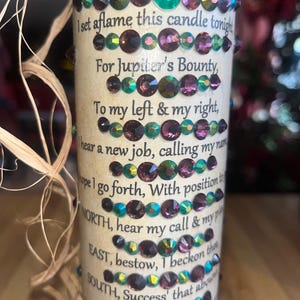 May include: A white candle with a brown label that reads "New Job Spell Candle". The label has a poem about finding a new job and includes the words "Jupiter's Bounty", "NORTH", "EAST", "SOUTH", and "WEST". The candle is decorated with iridescent green and purple rhinestones.