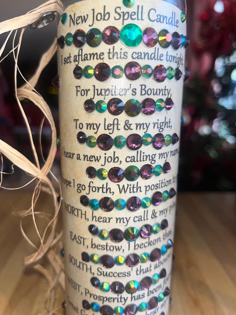 May include: A white candle with a brown label that reads "New Job Spell Candle". The label has a poem about finding a new job and includes the words "Jupiter's Bounty", "NORTH", "EAST", "SOUTH", and "WEST". The candle is decorated with iridescent green and purple rhinestones.