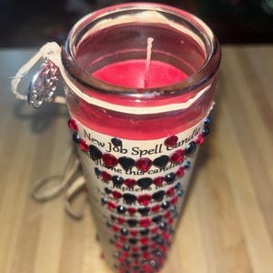 May include: A red candle with a white wick in a clear glass jar. The jar is decorated with black and red rhinestones and a label that reads "New Job Spell Candle".