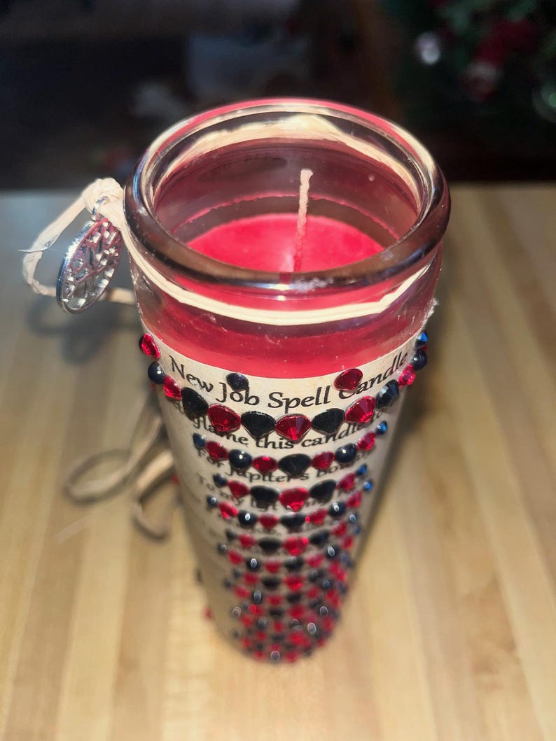 May include: A red candle with a white wick in a clear glass jar. The jar is decorated with black and red rhinestones and a label that reads "New Job Spell Candle".
