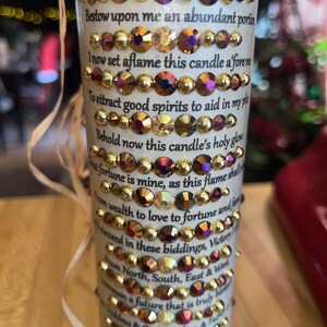 K&ouml;nnte beinhalten: Eine wei&szlig;e Kerze mit goldenen und roten Strasssteinen. Die Kerze tr&auml;gt eine Textinschrift, die lautet: "To the Powers of Jupiter, of Great Good Fortune. Bestow upon me an abundant portion. I now set aflame this candle afore me. To attract good spirits to aid in my plan. Behold now this candle's holy glow. Great fortune is mine, as this flame shall guide me. From wealth to love to fortune and fame. I shall succeed in these biddings, Victory is mine. From North, South, East & West. I summon a future that is truly blessed. By my intentions, answer my plea. Abundance is mine for all eternity."