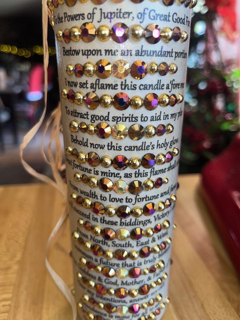 K&ouml;nnte beinhalten: Eine wei&szlig;e Kerze mit goldenen und roten Strasssteinen. Die Kerze tr&auml;gt eine Textinschrift, die lautet: "To the Powers of Jupiter, of Great Good Fortune. Bestow upon me an abundant portion. I now set aflame this candle afore me. To attract good spirits to aid in my plan. Behold now this candle's holy glow. Great fortune is mine, as this flame shall guide me. From wealth to love to fortune and fame. I shall succeed in these biddings, Victory is mine. From North, South, East & West. I summon a future that is truly blessed. By my intentions, answer my plea. Abundance is mine for all eternity."