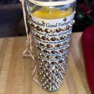 K&ouml;nnte beinhalten: Eine wei&szlig;e Kerze mit goldenen und schillernden Strasssteinen, mit dem Text "Abundant Good Fortune" und einem Gebet f&uuml;r Wohlstand.