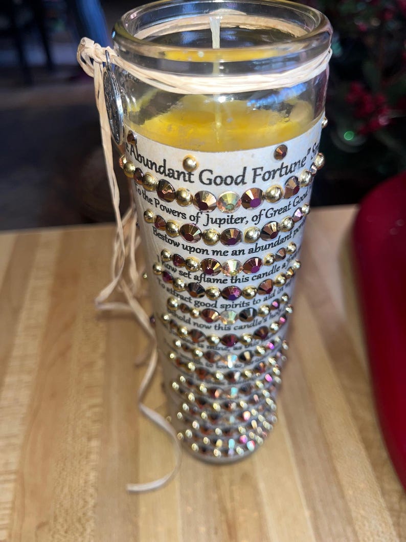 K&ouml;nnte beinhalten: Eine wei&szlig;e Kerze mit goldenen und schillernden Strasssteinen, mit dem Text "Abundant Good Fortune" und einem Gebet f&uuml;r Wohlstand.