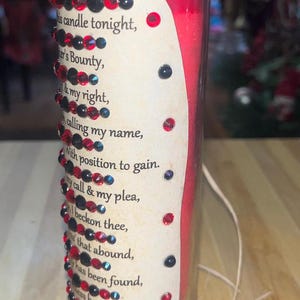 May include: A red glass candle with a white label that reads "Spell Candle" and a poem about calling upon a bounty. The candle is decorated with red and black rhinestones.