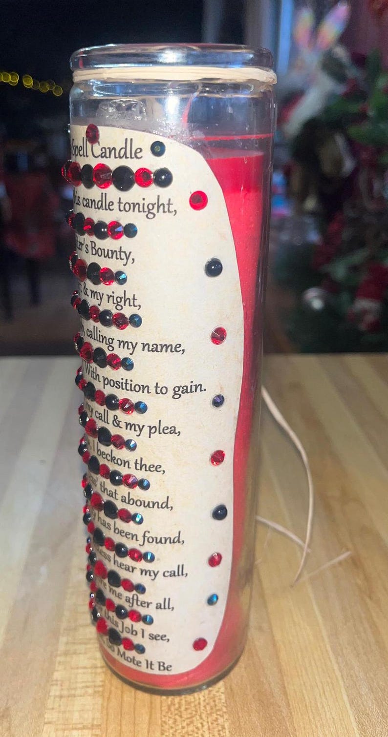 May include: A red glass candle with a white label that reads "Spell Candle" and a poem about calling upon a bounty. The candle is decorated with red and black rhinestones.