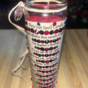 May include: A red candle with a beige label that reads "New Job Spell Candle" and has a decorative trim of red and black rhinestones. The candle is set on a wooden surface.
