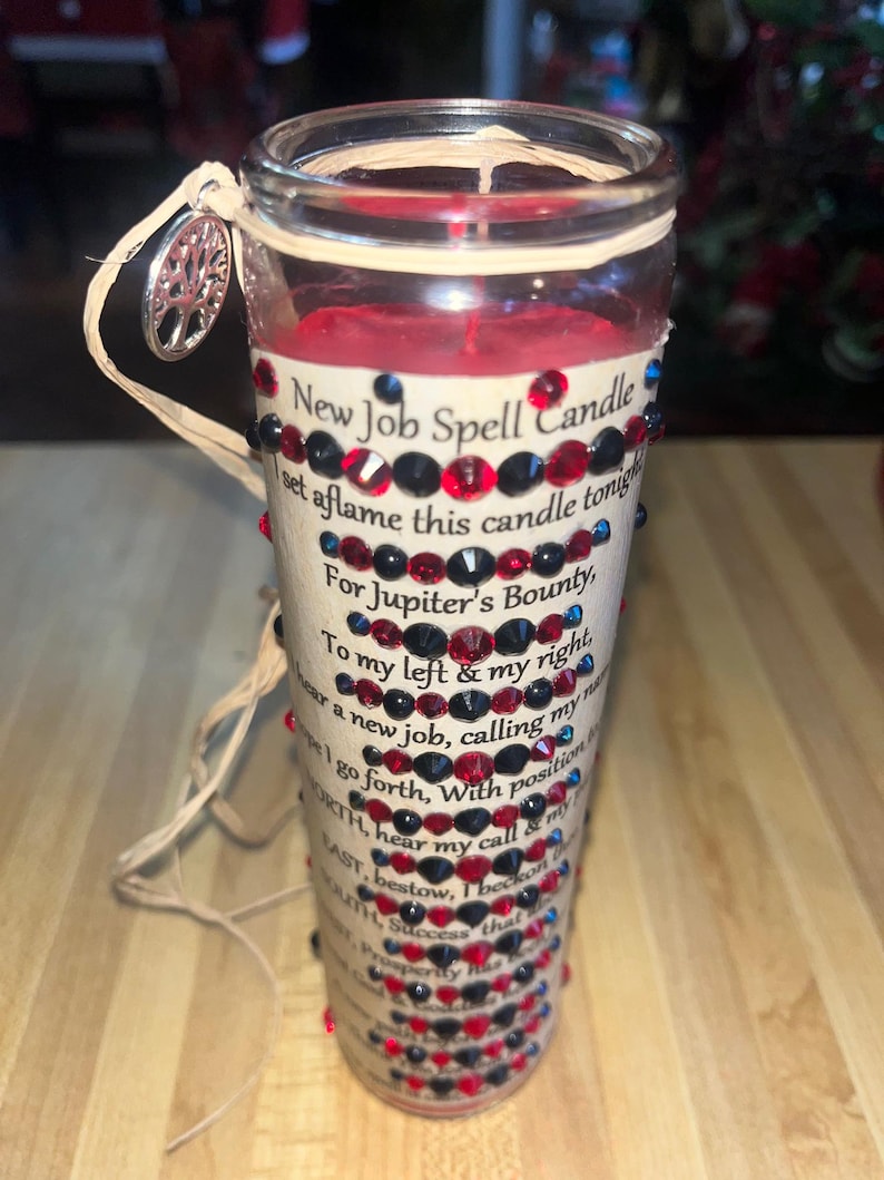 May include: A red candle with a beige label that reads "New Job Spell Candle" and has a decorative trim of red and black rhinestones. The candle is set on a wooden surface.