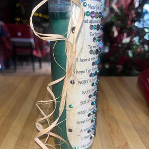 May include: Green glass candle with a tan ribbon wrapped around it. The candle has a label with the text "New Job" and a list of directions for a spell. The label is decorated with iridescent rhinestones.