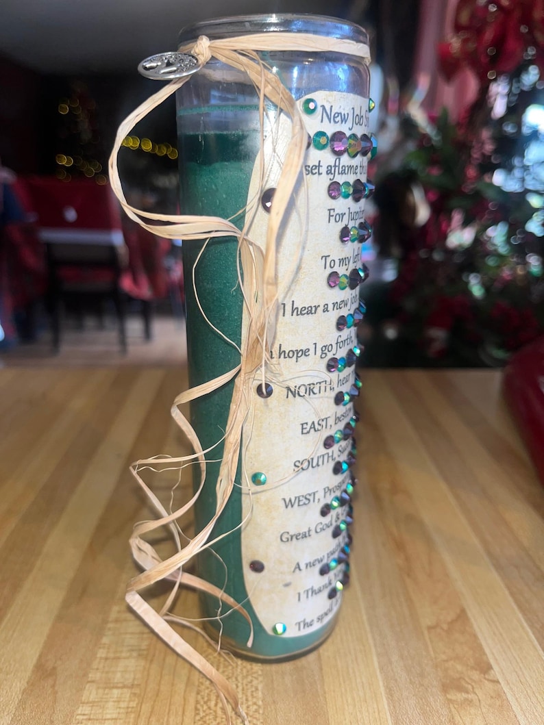May include: Green glass candle with a tan ribbon wrapped around it. The candle has a label with the text "New Job" and a list of directions for a spell. The label is decorated with iridescent rhinestones.