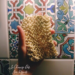 Könnte beinhalten: Nahaufnahme einer Hand, die einen hellbraunen, gestrickten Luffa hält. Der Luffa besteht aus natürlichen Fasern und hat eine raue Textur. Der Hintergrund ist eine gekachelte Wand mit einem bunten geometrischen Muster. Der Text "Le Champ Des Sens Reines" ist in der linken unteren Ecke des Bildes sichtbar.