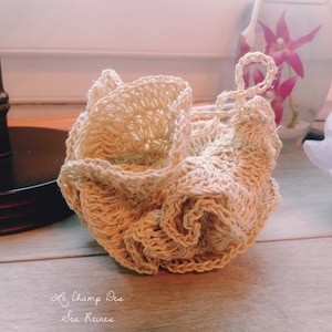 Può includere: Un pouf da bagno o uno scrub in crochet beige con un manico annodato. Il pouf è fatto di fibre naturali e ha una superficie testurizzata. Il nome del marchio "Le Champ Des Sea Reines" è scritto in corsivo nella parte inferiore dell'immagine.