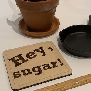 Peut inclure: Une pancarte en bois avec le texte "Hey, sugar!" en lettres brunes. La pancarte est sur une surface blanche avec une plante en pot et une poêle en fonte.