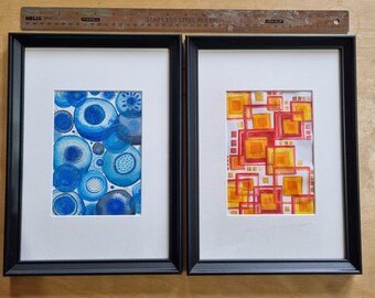 May include: Two framed abstract watercolor paintings. The painting on the left features a blue and white pattern of overlapping circles. The painting on the right features a red and yellow pattern of overlapping squares.