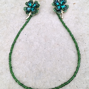 Puede incluir: Un par de pendientes verdes con forma de trébol con piedras preciosas de color esmeralda. Los pendientes están conectados por un hilo de pequeñas cuentas verdes. El trébol tiene un tallo dorado.