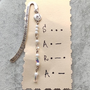 Op de afbeelding: Een zilveren boekenlegger met een decoratief wervelontwerp en een kralenbedel. De bedel bevat witte kralen en iriserende kristallen. Een kleine kaart met de naam "SARA" in morsecode is bevestigd.