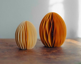 May include: Two paper honeycomb Easter eggs, one yellow and one orange, on a wooden surface.
